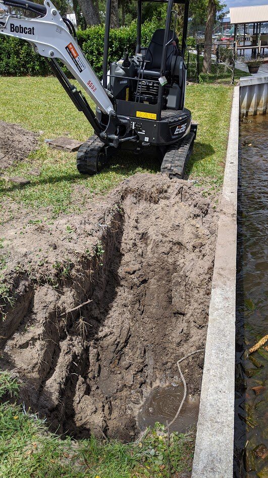 Compact Bobcat mini-excavator working in tight backyard access excavating along the same St. Johns River seawall repair job